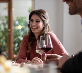 Donna sorridente che conversa con un uomo mentre beve un bicchiere di vino in un ambiente accogliente.