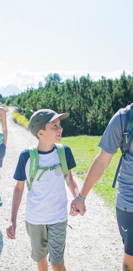 Famiglia sorridente in escursione su un sentiero soleggiato con staccionata e paesaggio verde.