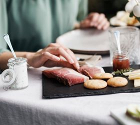 Una persona prende una fetta di speck da un tagliere con specialità altoatesine come formaggio, pane, quark alle erbe e marmellata.