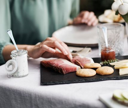 Una persona prende una fetta di speck da un tagliere con specialità altoatesine come formaggio, pane, quark alle erbe e marmellata.