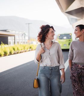 Due donne chiacchierano sorridendo a una fermata dell’autobus moderna con un autobus verde sullo sfondo.