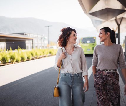 Due donne chiacchierano sorridendo a una fermata dell’autobus moderna con un autobus verde sullo sfondo.