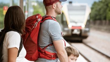 Famiglia con zaini aspetta il treno in arrivo sul marciapiede.