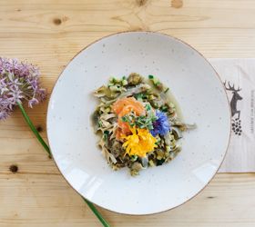 Piatto con pasta, verdure, salmone e fiori edibili su un tavolo di legno chiaro con un fiore.