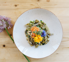 Piatto con pasta, verdure, salmone e fiori edibili su un tavolo di legno chiaro con un fiore.