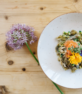 Piatto con pasta, verdure, salmone e fiori edibili su un tavolo di legno chiaro con un fiore.