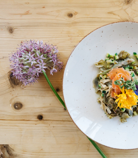 Piatto con pasta, verdure, salmone e fiori edibili su un tavolo di legno chiaro con un fiore.