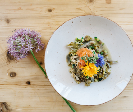 Piatto con pasta, verdure, salmone e fiori edibili su un tavolo di legno chiaro con un fiore.